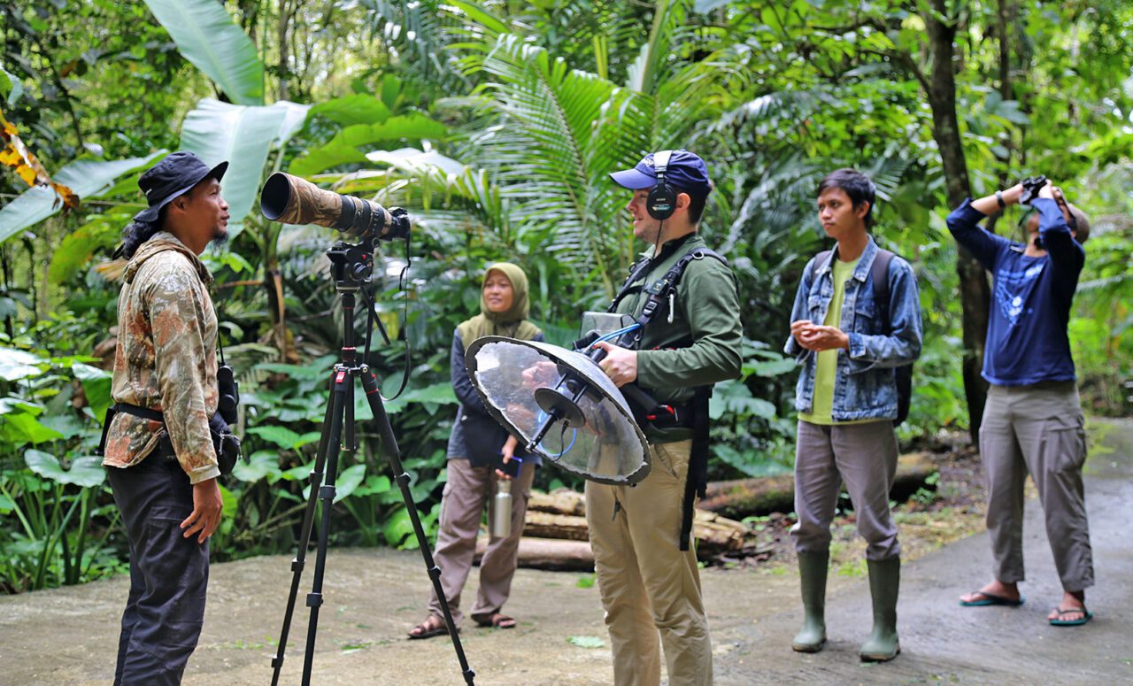 A group with binoculars, cameras and audio equipment, stands in a forest.