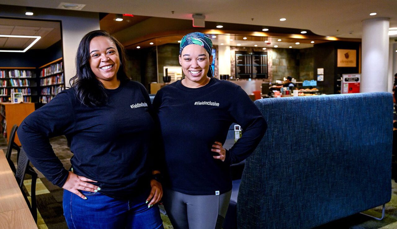 Two women stand in a bookstore and smile at the camera with Tshorts that say #FieldInclusive