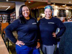 Two women stand in a bookstore and smile at the camera with Tshorts that say #FieldInclusive