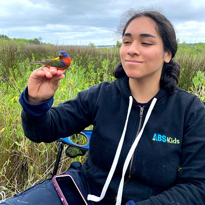 A woman with a blue sweatshirt, holds a small, rainbow colored bird