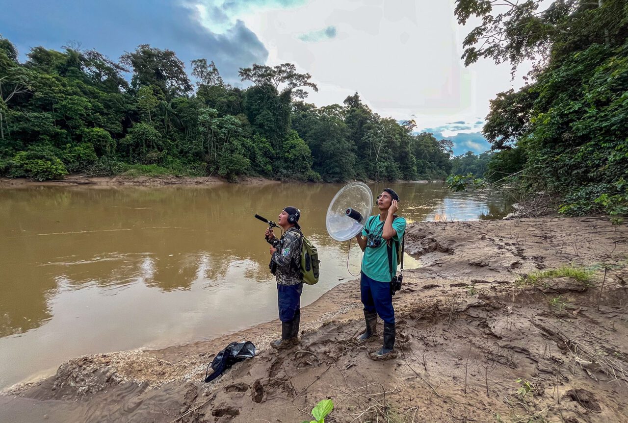 Two men stand by a forested river with sound recording equipment.