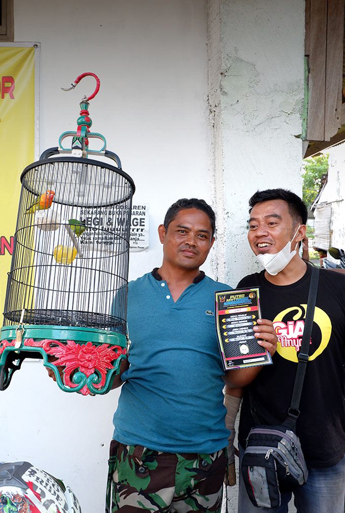 Two man stand together, one holds a bird cage with two birds inside.