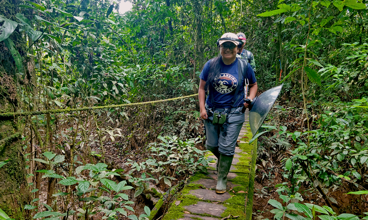 A woman walks in the forest with sound recording equipment.